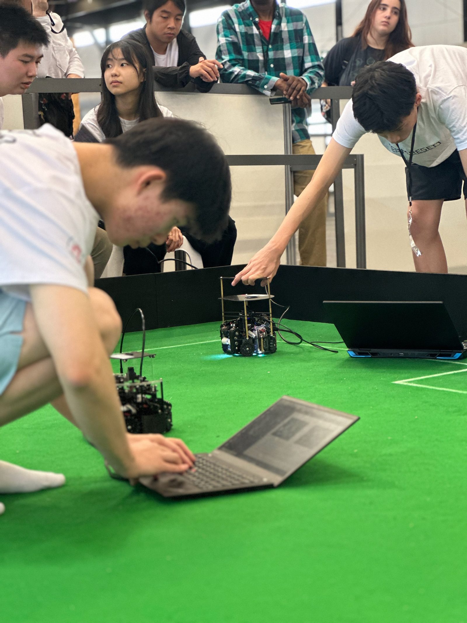 RoboCupJunior Soccer: estudiantes en cuclillas alrededor de una cancha verde mientras pequeños robots autónomos juegan; un competidor ajusta el código en una laptop a un costado de la cancha.